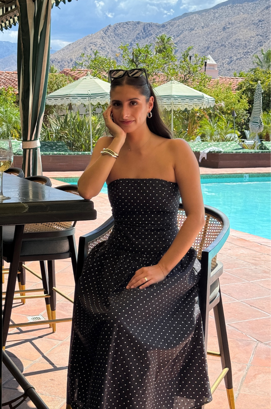 Woman in a black polka dot dress sitting by a poolside with mountains in the background