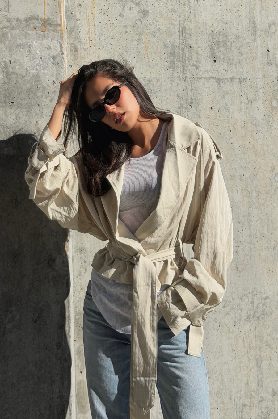 Woman wearing a beige trench coat over a white top and blue jeans against a concrete wall.