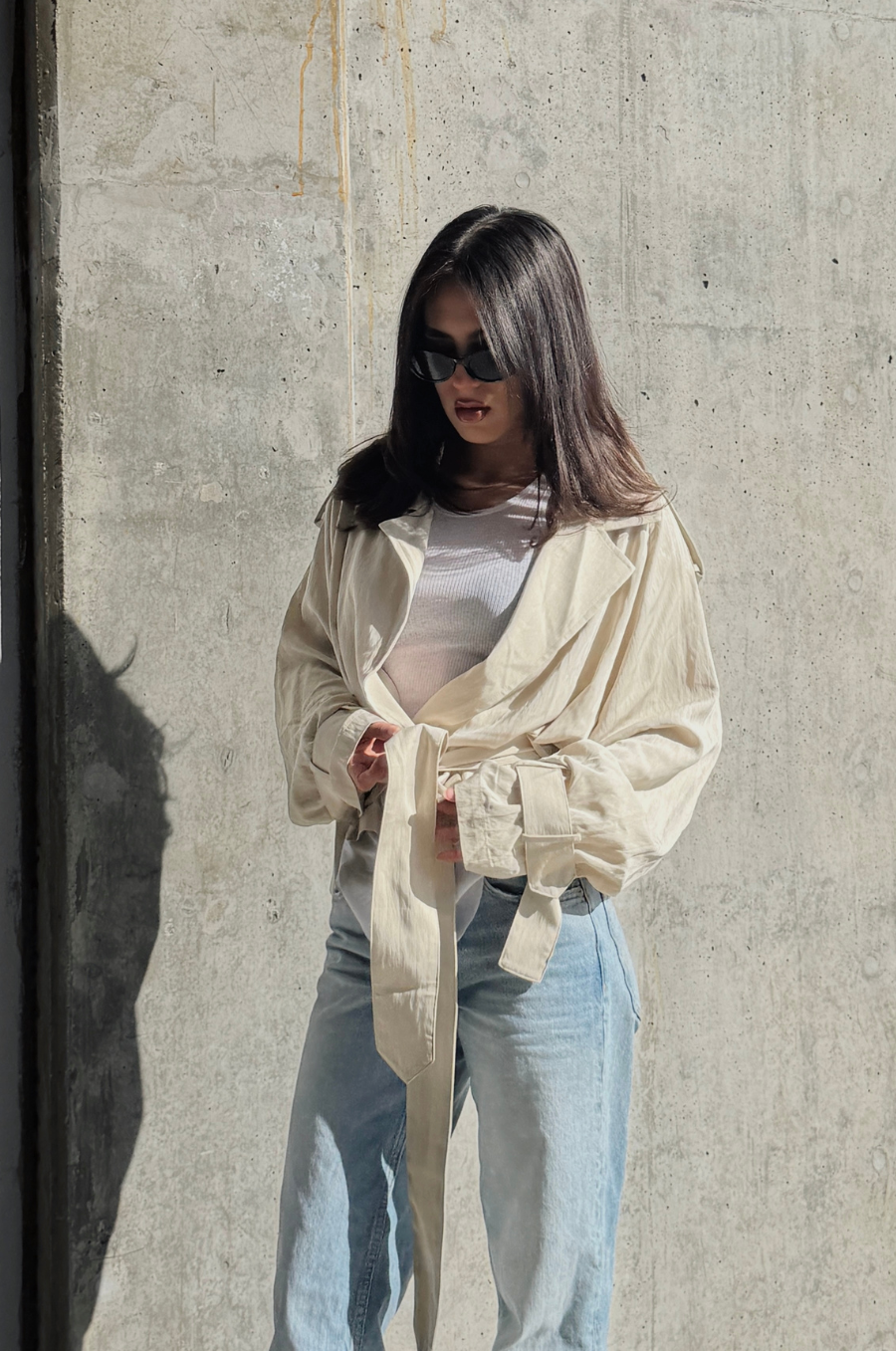 Woman wearing a cream-colored shirt and light blue jeans against a concrete wall.