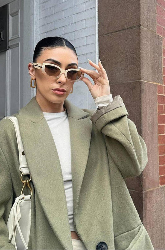 Woman wearing a green coat and sunglasses, standing against a brick wall.