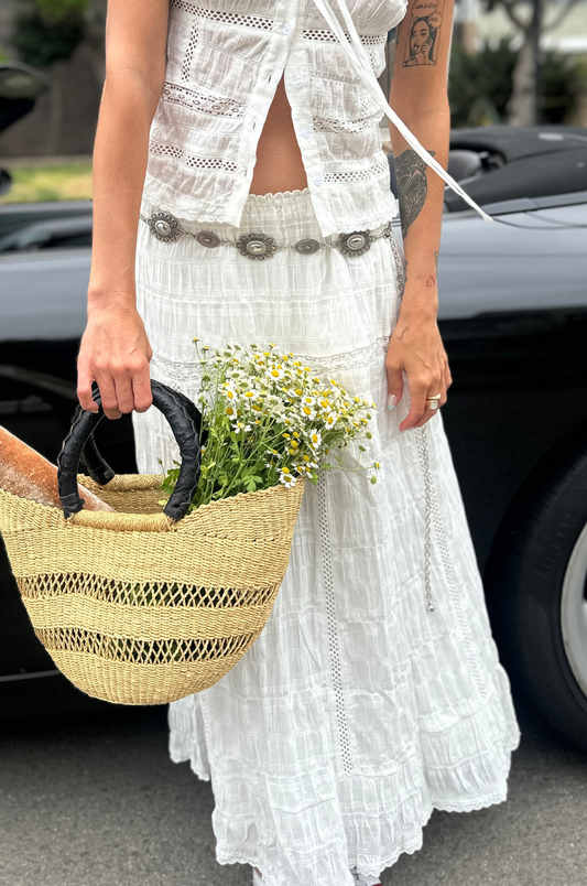Person wearing a white dress holding a woven bag with flowers, standing next to a car.