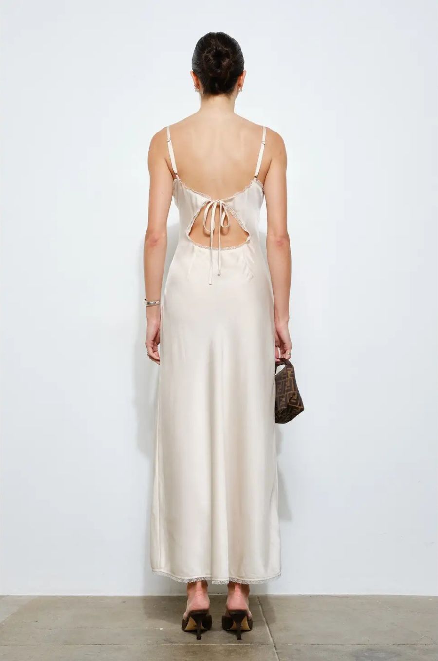 Woman wearing a white satin dress with thin straps and open back, holding a small black handbag.