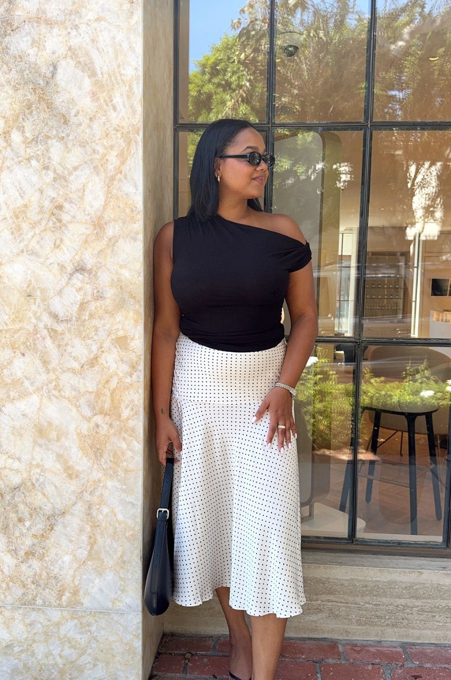 Woman in black top and white skirt standing against a textured wall with glass doors.